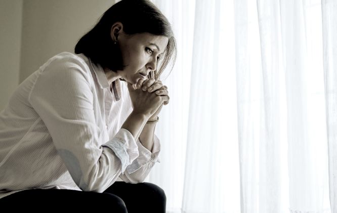 A person sitting pensively with hands clasped near the mouth, staring into the distance, by a window with sheer curtains, contemplating the steps ahead in their divorce mediation.